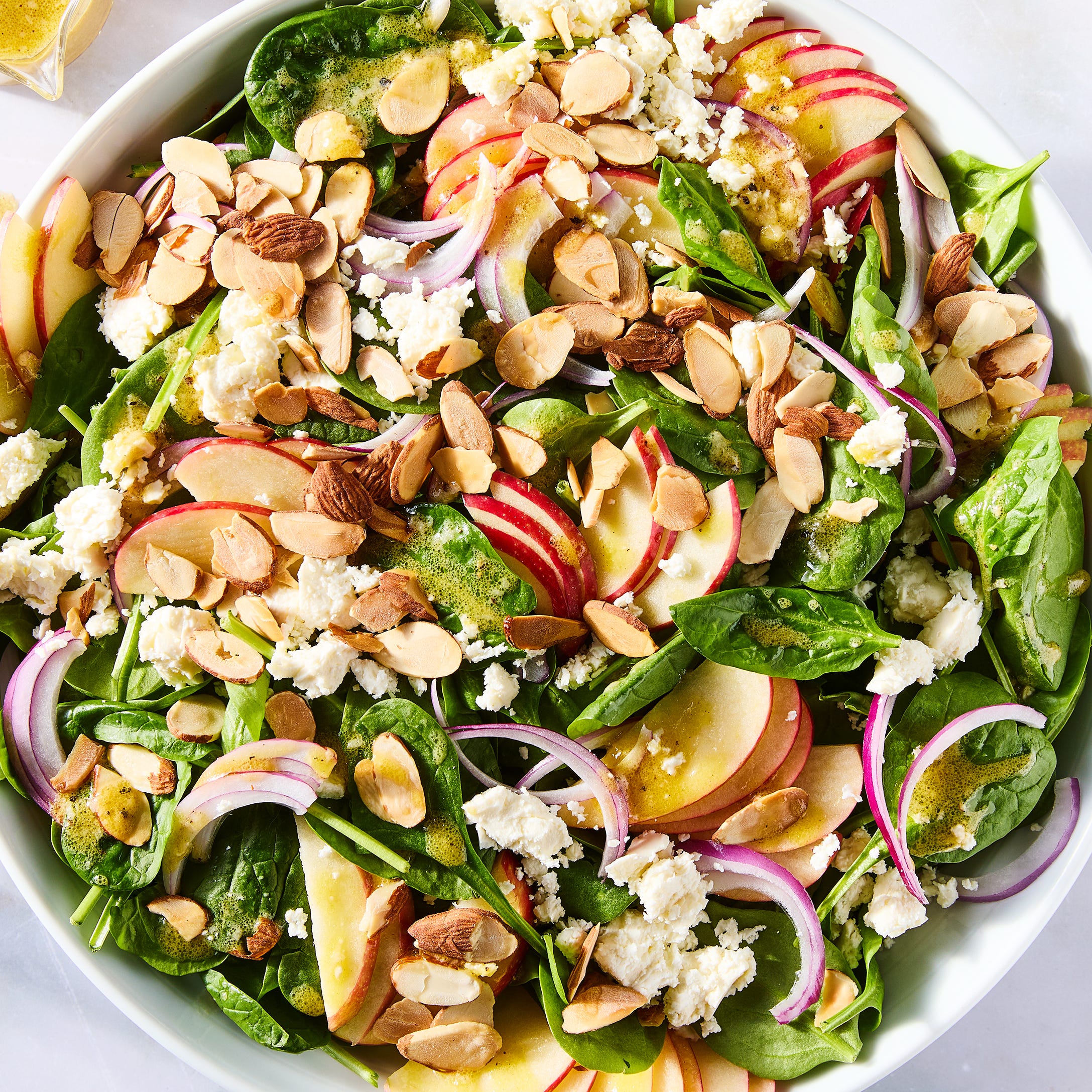 spinach in a bowl topped with sliced apples, sliced almonds, red onions, and crumbled feta