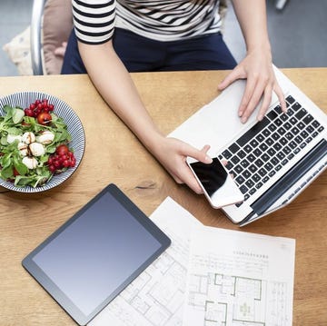 Woman at desk using laptop and cell phone next to construction plan and salad woman at desk using laptop and cell phone next to construction plan and salad