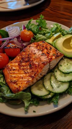 a white plate topped with salmon, cucumber and tomatoes next to lettuce