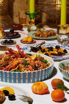 A spread of Moroccan dishes on a table. In the foreground, peeled clementines and a prawn tagine in a blue and white serving dish. In the background, a citrus and olive salad, goat cheese cigars, a dish of kumquats, long candlesticks, and Moroccan lanterns.