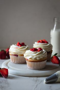 three cupcakes with white frosting and strawberries on a plate next to a bottle of milk