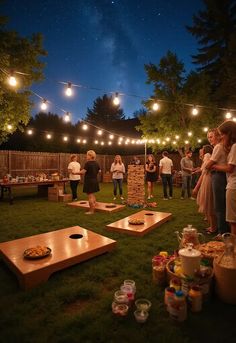 a group of people standing around in the grass with lights strung over them and food on picnic tables