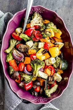 a purple bowl filled with roasted vegetables on top of a gray table cloth next to a fork