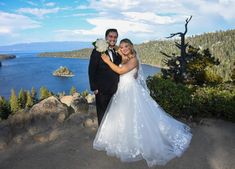 a bride and groom pose for a photo on top of a mountain overlooking the lake