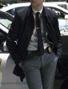 a man in a suit and tie standing next to a police car with his hands in his pockets
