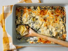 a casserole dish with spinach and cheese on top, next to a wooden spoon