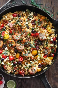 a skillet filled with pasta and vegetables on top of a wooden table next to olives