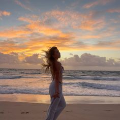 a woman standing on top of a sandy beach next to the ocean at sunset with her hair blowing in the wind
