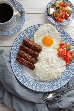 a blue and white plate topped with rice, sausages and an egg next to a cup of coffee