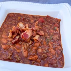 a white square bowl filled with beans and bacon in a chili based sauce on top of a blue table cloth