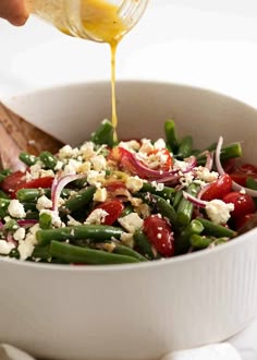 someone pouring dressing into a white bowl filled with green beans, tomatoes and feta cheese