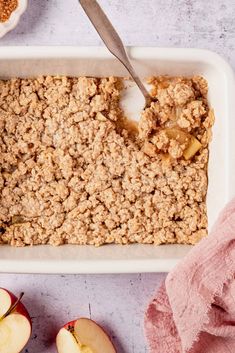 an apple crisp in a white baking dish with a spoon next to it and two apples on the side