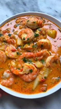 a white bowl filled with soup and shrimp on top of a marble counter next to a hand holding a spoon
