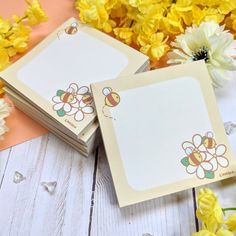 two cards with bees on them sitting next to yellow and white flowers in the background