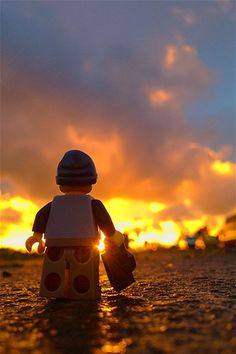 a lego figure standing in the middle of a street at sunset with clouds and sun behind it