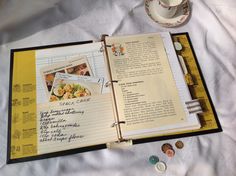 an open recipe book sitting on top of a bed next to a cup and saucer