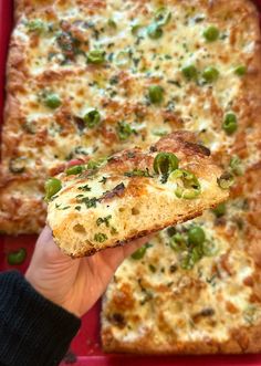 a person holding up a piece of pizza in front of a red tray with green toppings