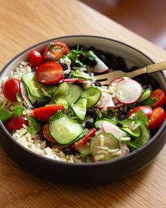 a bowl filled with rice, cucumbers, tomatoes and black olives next to a wooden table