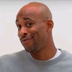 a bald man wearing a gray sweater and smiling at the camera with his eyes wide open