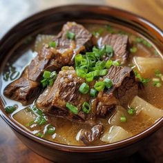 a brown bowl filled with beef and potatoes on top of a wooden table next to green onions