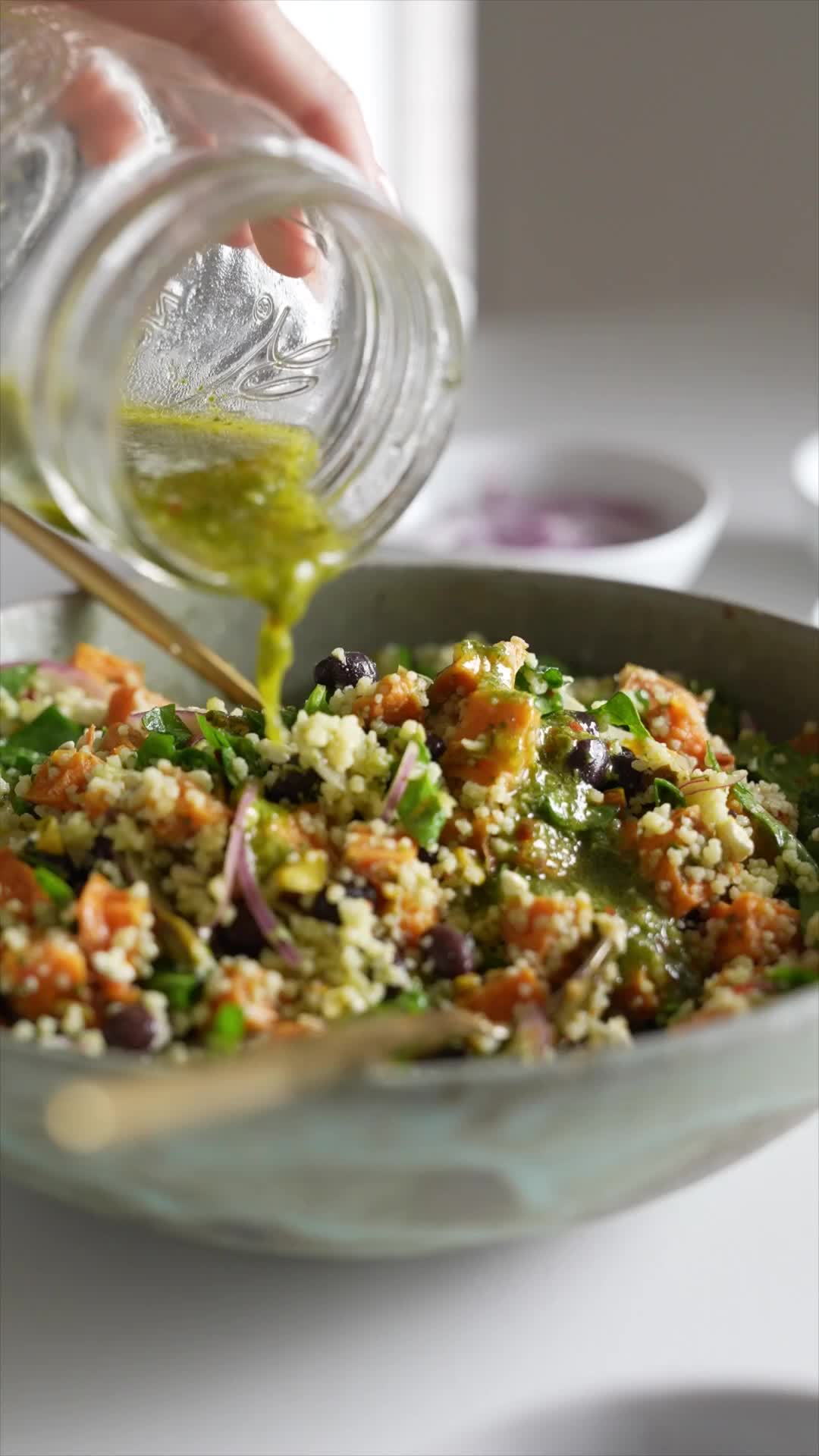 This contains: Couscous is added to a bowl along with the other salad ingredients - cooked cubed sweet potatoes, chopped spinach, feta cheese, black beans, red onion, and chopped pistachios. A lime basil vinaigrette is poured on top and the whole salad is tossed together.