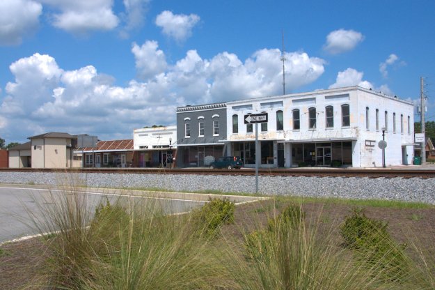 A view of a small town street featuring historic buildings, a railway track in front, and a 'One Way' sign under a partly cloudy sky.