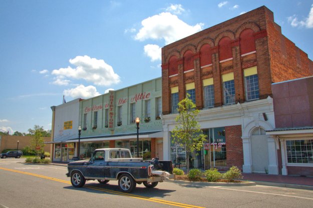 A street view of a small town featuring two storefronts, one with a sign reading 'Clothes in the Attic Thrift Store' and another building with large windows and decorative elements. A blue pickup truck is parked in front, and the scene is set under a bright sky with fluffy clouds.