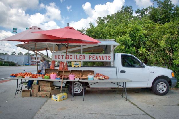 A roadside stand selling hot boiled peanuts and tomatoes under a red umbrella, with a white pickup truck parked nearby.