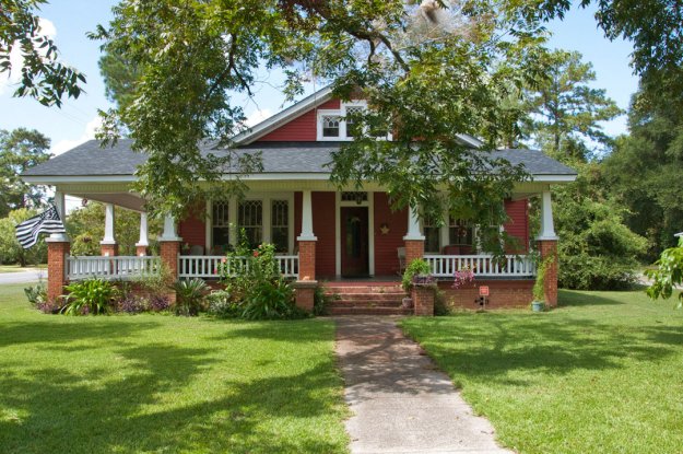A charming red Craftsman cottage with a front porch surrounded by green grass and plants, featuring white railings and brick columns.