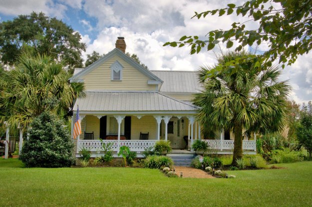 A charming yellow house with a metal roof, featuring a white porch with rocking chairs and an American flag, surrounded by lush greenery and palm trees under a partly cloudy sky.