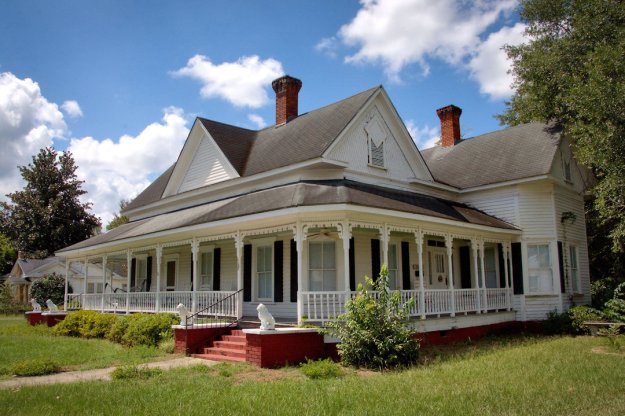 Historic Victorian house with a large porch, black shutters, and red brick steps, surrounded by green grass and trees under a partly cloudy sky.