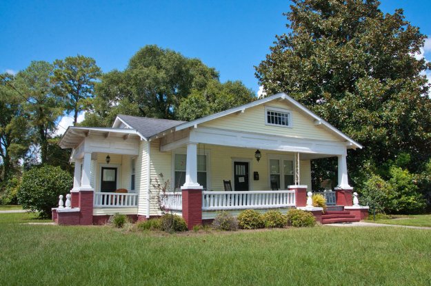 A charming yellow bungalow house with a front porch, white columns, and red brick accents, surrounded by a green lawn and trees.