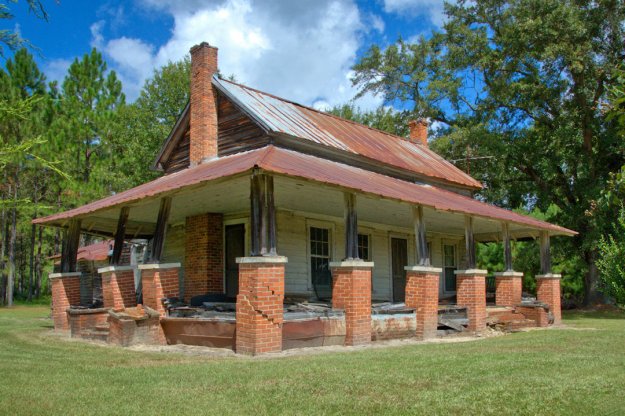 An abandoned historic house with a rusty metal roof, brick columns, and a weathered wooden structure surrounded by lush greenery.