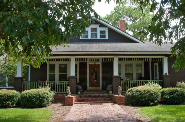 Front view of a brown wooden house with a gabled roof, featuring a porch with white columns, surrounded by greenery and shrubs.