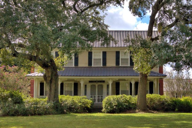A classic two-story house with a metal roof, surrounded by trees and bushes, showcasing a traditional architectural style.