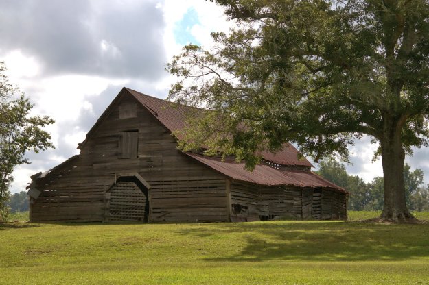 An old wooden barn with a red roof, surrounded by green grass and trees, under a partly cloudy sky.