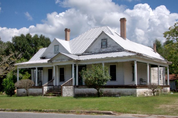 A weathered, two-story house with a metal roof and front porch, surrounded by greenery and under a partly cloudy sky.
