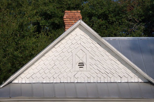 View of a gable roof featuring white wooden shingles and a brick chimney, with a decorative vent in the center.