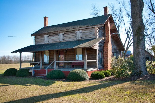 A rustic wooden house with a porch, featuring brick chimneys and a sloped roof, surrounded by manicured bushes on a sunny day.