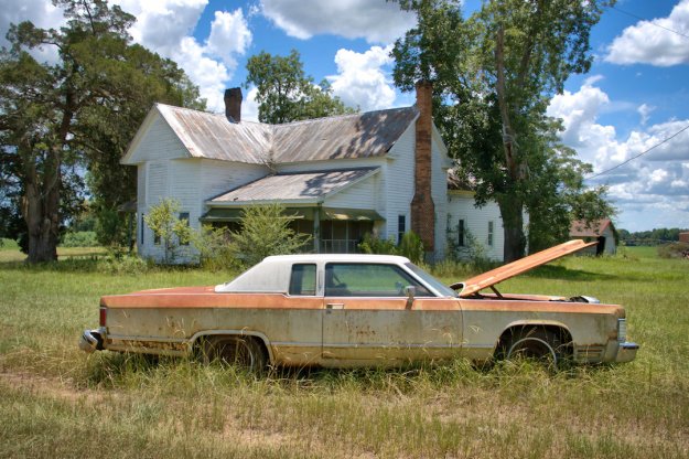 An old, rusty Lincoln car with its hood open parked in tall grass in front of a dilapidated white house under a cloudy blue sky.