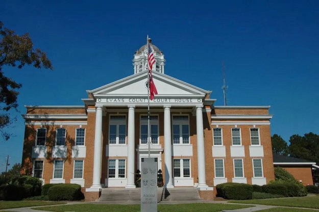Front view of Evans County Court House with a blue sky, featuring American and state flags, and landscaped yard.