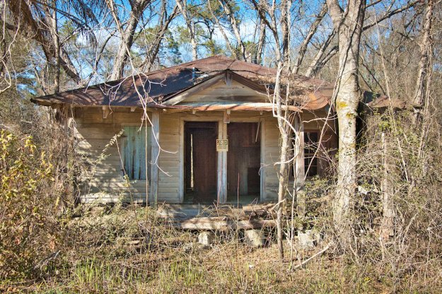 Abandoned wooden house surrounded by overgrown vegetation, featuring a rusty roof and double front doors.