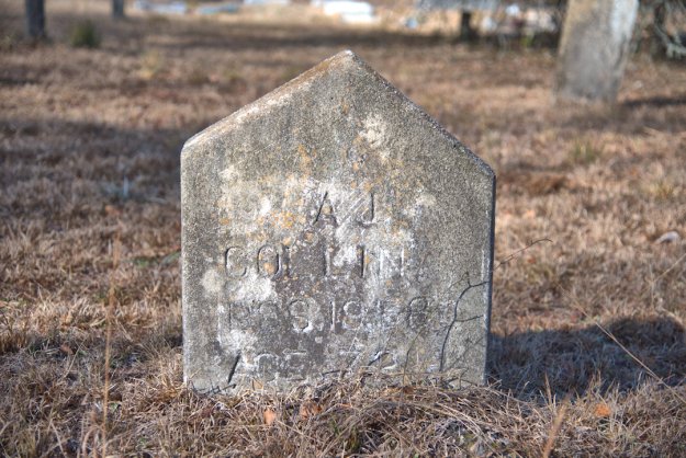 A weathered gravestone in a cemetery, engraved with the name 'A.J. Collins' along with dates and age information, surrounded by dry grass.
