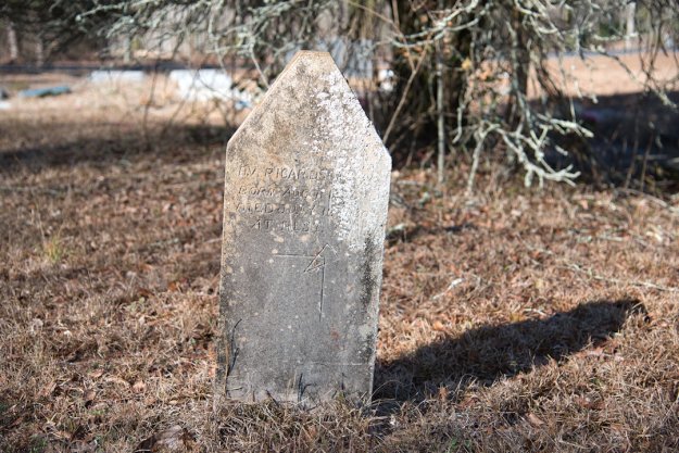 A weathered grave marker in a cemetery, partially covered in moss, with inscriptions indicating the name, birth date, and death date of the individual it commemorates.