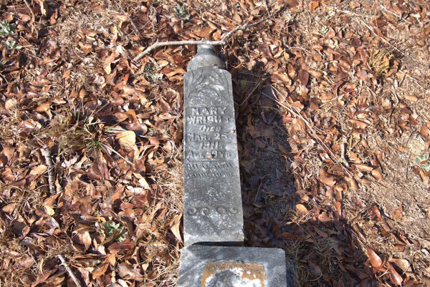 A weathered gravestone engraved with the name 'Mary Wright,' the inscription 'Died Mar. 22, 1911,' and 'Aged 79.' The stone is surrounded by dry leaves and grass.
