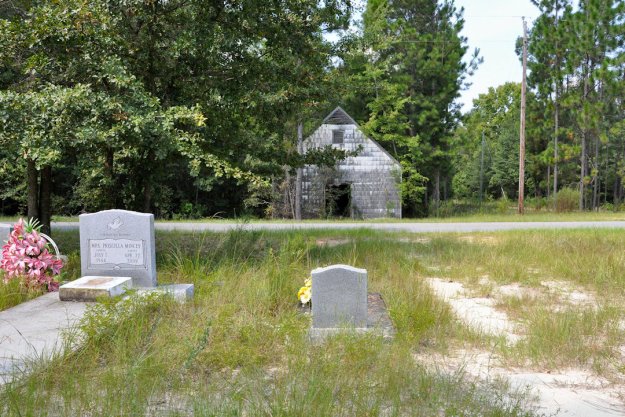 Gravestones in a grassy area with a dilapidated building in the background surrounded by trees.