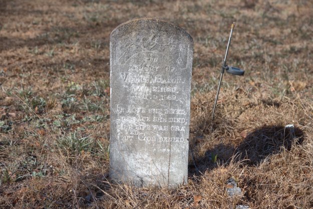 A weathered gravestone in a cemetery, inscribed in memory of Venus Bacon, with dates of birth and death visible, surrounded by dry grass.