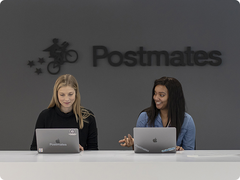 The image shows two individuals seated at a white desk, each working on a laptop. The person on the left has long blonde hair and is wearing a black hoodie, while the person on the right has long dark hair and is wearing a blue shirt. Both laptops have stickers on them. Behind them, there is a large logo of "Postmates" with an image of a person riding a bicycle and stars trailing behind.