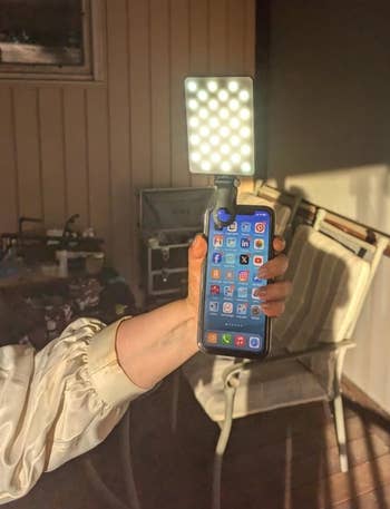 Person holding a smartphone with apps visible, attached to a square LED light. Background shows a porch with a chair and table