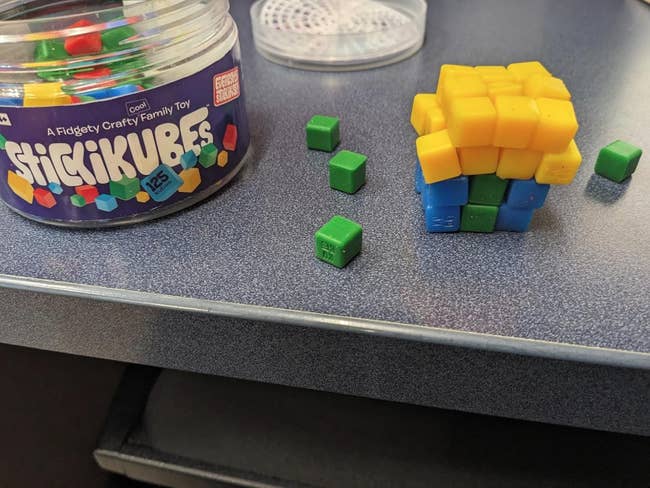 StickiKubes toy jar beside partially assembled cube made of green and yellow pieces on a desk